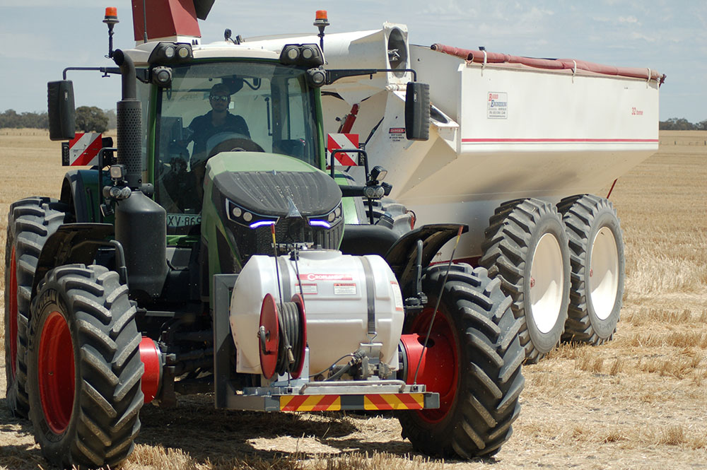 A green tractor with a fire fighting pump and water tank on the front is towing a white metal chaser bin during wheat harvesting.