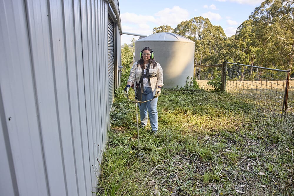  A woman using a brushcutter to clear long grass beside a shed, wearing protective earmuffs, gloves, and safety gear, with a water tank and trees in the background.