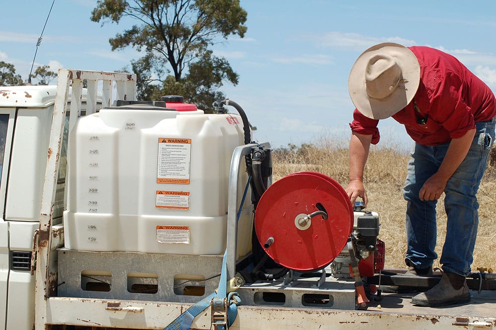 a farmer in a red shirt, blue jeans and a beige felt hat stands on the back of a ute, which has a simple slip-on on the tray, made up of a fire fighting hose, pump and a couple of hundred litres of water.