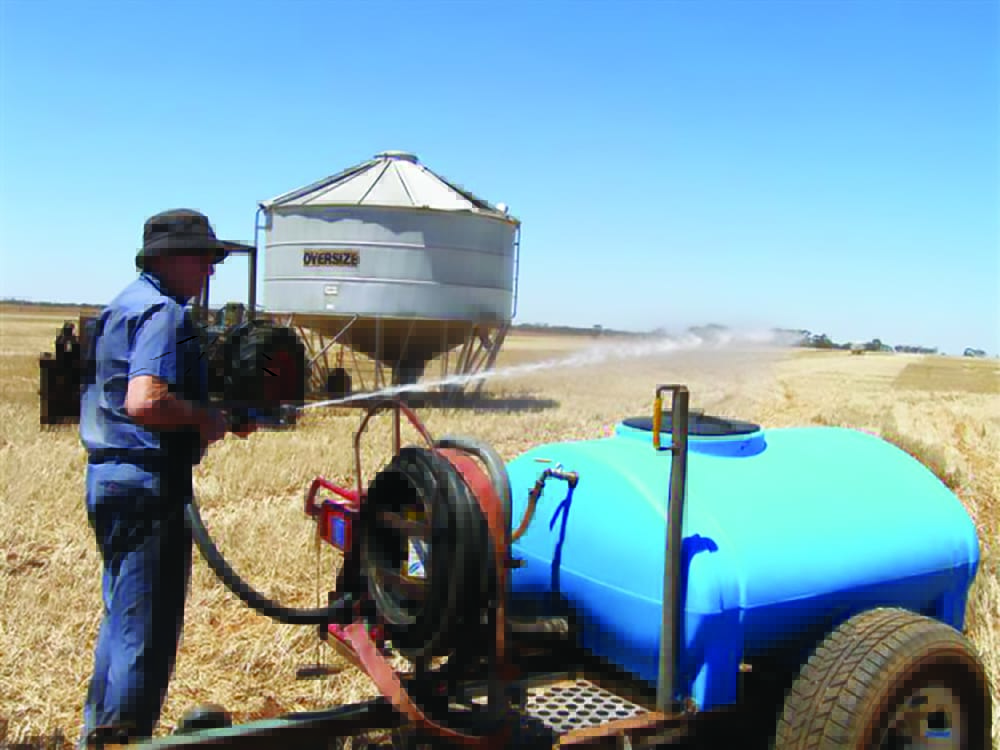 A farmer stands in a harvested wheat field holding a fire hose which is attached to a water tank and pump on a trailer.