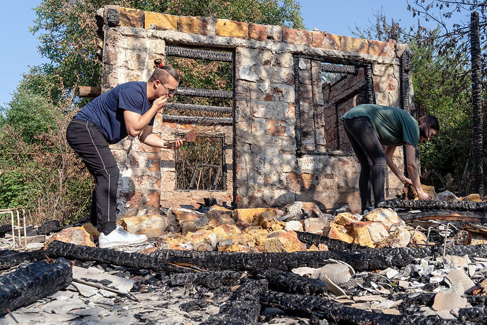 Two people sifting through charred rubble of a burned building, with a partially standing stone wall behind them.