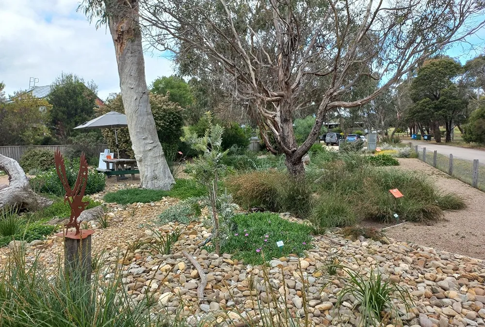 Low growing groundcover plants, native grasses and other plants are broken up by stone mulch and a dry creek bed. There is picnic table and chairs in an area with crushed rock on the ground.
