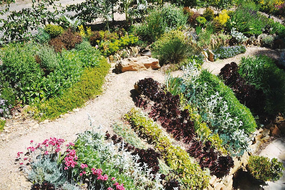 A number of garden beds with colourful and green low lying plants and shrubs are broken up with gravel paths and rockery.