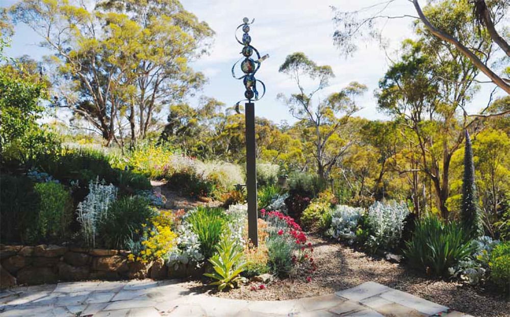 Sandstone paving, river pebbles and gravel separate garden beds containing colourful low lying and low flammability plants. In the distance is a native forest.
