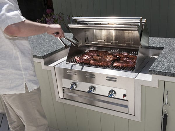 A person using tongs and cooking steaks on a built-in outdoor gas BBQ.