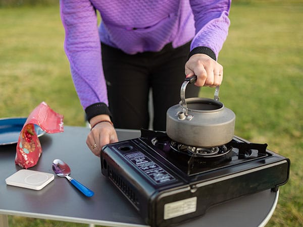 A person heating a kettle using a portable butane gas stove which is sitting on a level table.