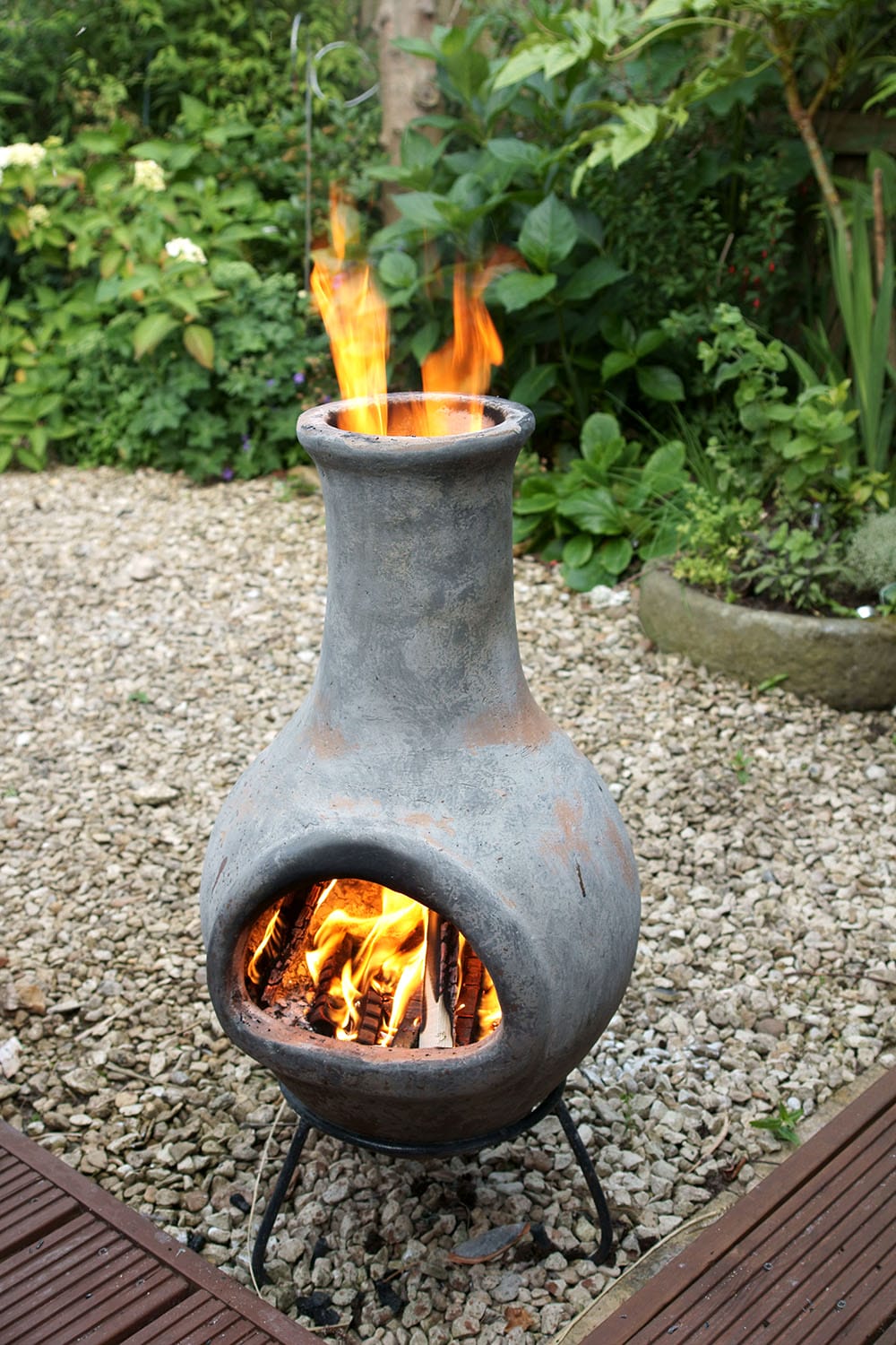 A bulb-shaped chimenea full of small burning logs sitting in a metal frame. Flames are coming out of the chimney at the top. It has been put on a gravel (?) area next to a deck and there are shrubs in the background.