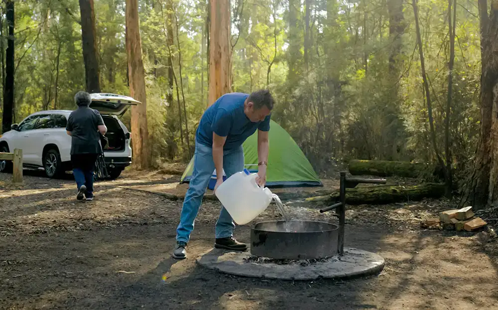 A woman carries a camp chair toward an open car in a bushland camp site. A man is in the foreground pouring water from a large plastic container onto a campfire. There is a green tent behind the man and tall trees and scrub in the background.