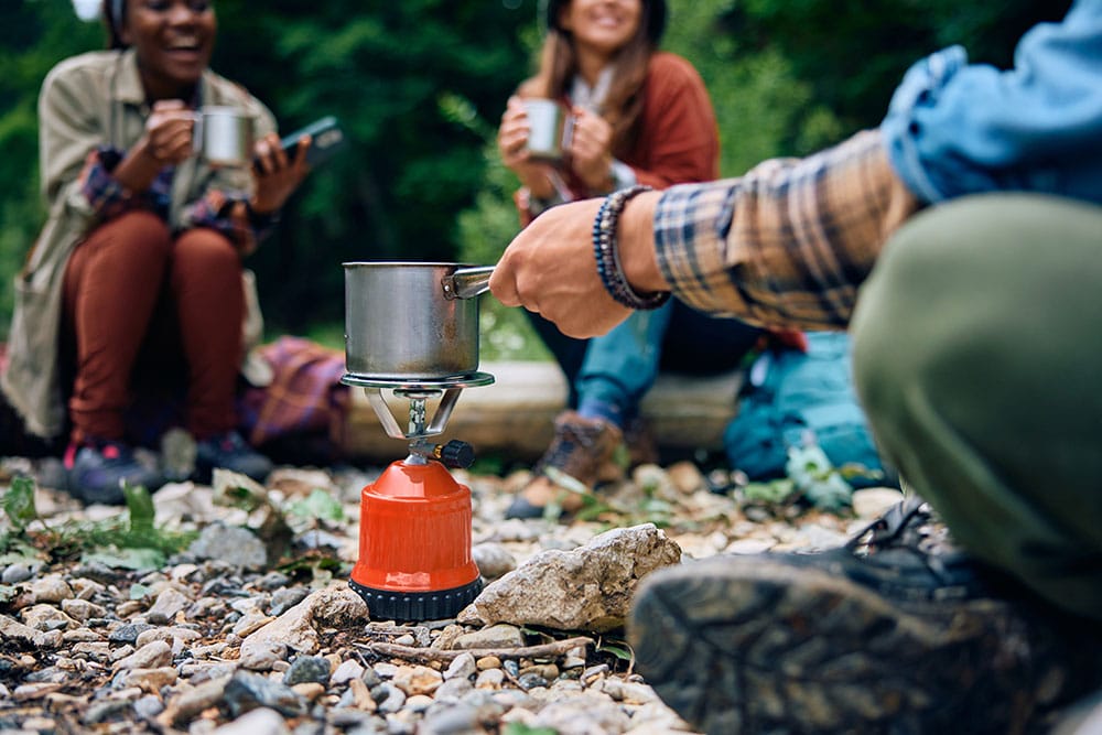 Hiker heating a metal pot on a stable portable gas stove placed on a cleared rocky forest floor, with two hikers sitting in the background holding metal mugs.