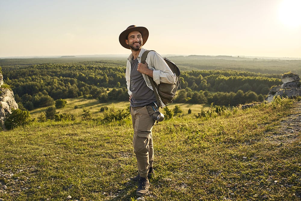 Hiker wearing a wide-brimmed sun hat, lightweight long-sleeve hiking shirt layered over a t-shirt, and tan cargo pants, carrying a durable backpack with an attached metal water bottle and sturdy hiking boots, standing on a mountain overlooking dry, bushfire-prone terrain.