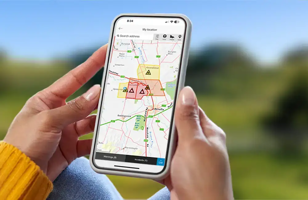 Close up of a woman's hands holding a phone showing a map with coloured shapes representing areas affected by fire. In the background can be seen out of focus rolling hills and trees.