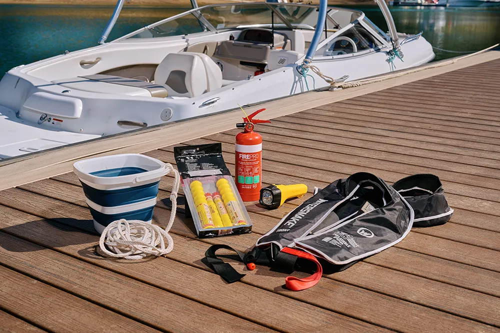 White speed boat moored at a dock with boating safety essentials including a collapsible bucket with rope attached, flares, fire extinguisher, floating torch, and life jacket on the wooden deck.