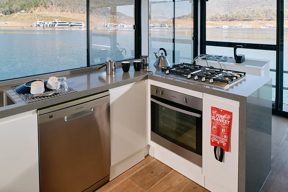 The kitchen on a houseboat with a lake scene in the background with other houseboats in the distance. In the kitchen is a sink, gas cooktop, oven and coffee making facilities. A fire blanket hangs near the stove.