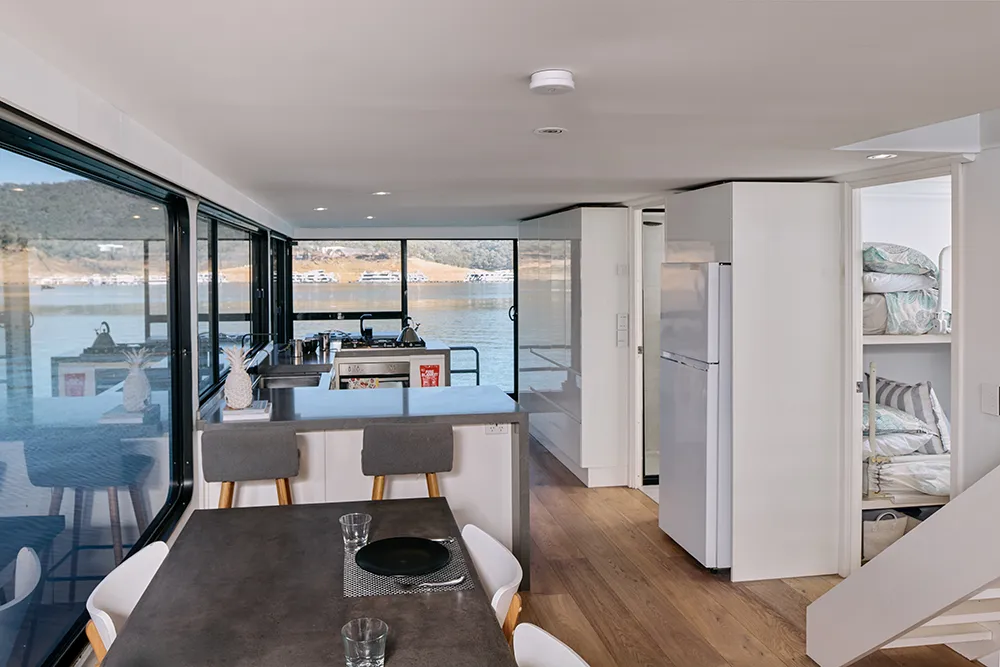 The interior of a houseboat with a view of a lake outside. A set dining table is in the foreground and behind that is a kitchen area with sink, stove, kettle and refrigerator. To the right is the open door to a bedroom and a smoke alarm is on the ceiling outside the bedroom.