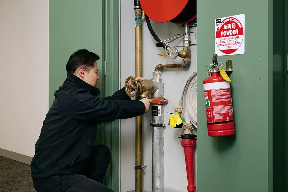 A person wearing CFA uniform inspects a fire hose reel and valve in a utility cupboard. Installed on cupboard door is a red fire extinguisher with a sign above labelled "A:B(E) Powder".