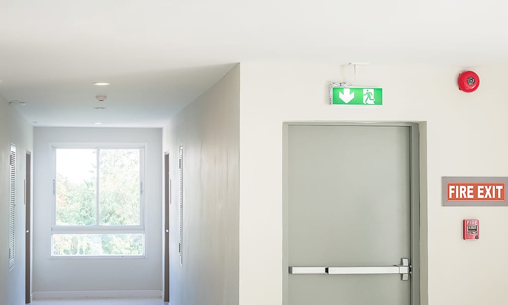An exit sign is illuminated above a fire door in a building. There is an pointing down next to an icon of a person running through a door on the sign.