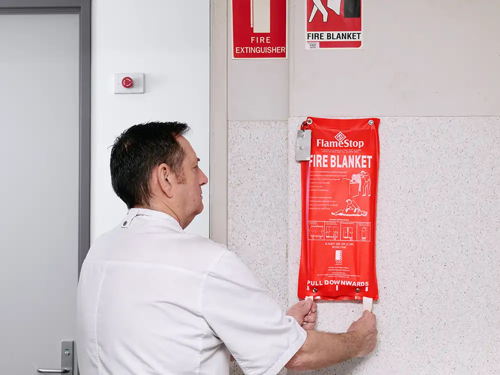 A chef in a commercial kitchen pulling the tabs to remove a fire blanket from its cover.