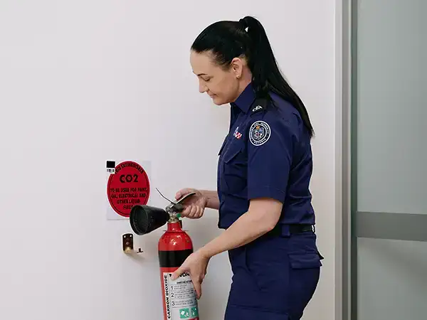 A person in a CFA uniform is taking a fire extinguisher off a wall. Above the fire extinguisher is a square plastic sign. There is a red circle on the sign with the words, in black: 'This extinguisher: CO2, to be used for paint, oil, electrical and other liquid fires.'