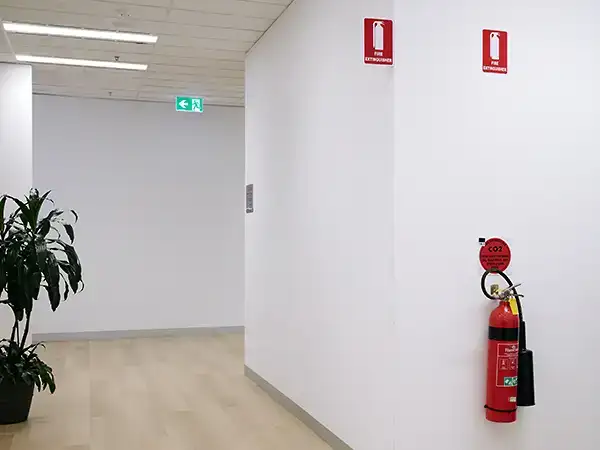 Signs attached to the top of either side of a corner of a hallway wall. They are red background with a white icon of a fire extinguisher and the words 'fire extinguisher'. A fire extinguisher is located directly under one of the signs.