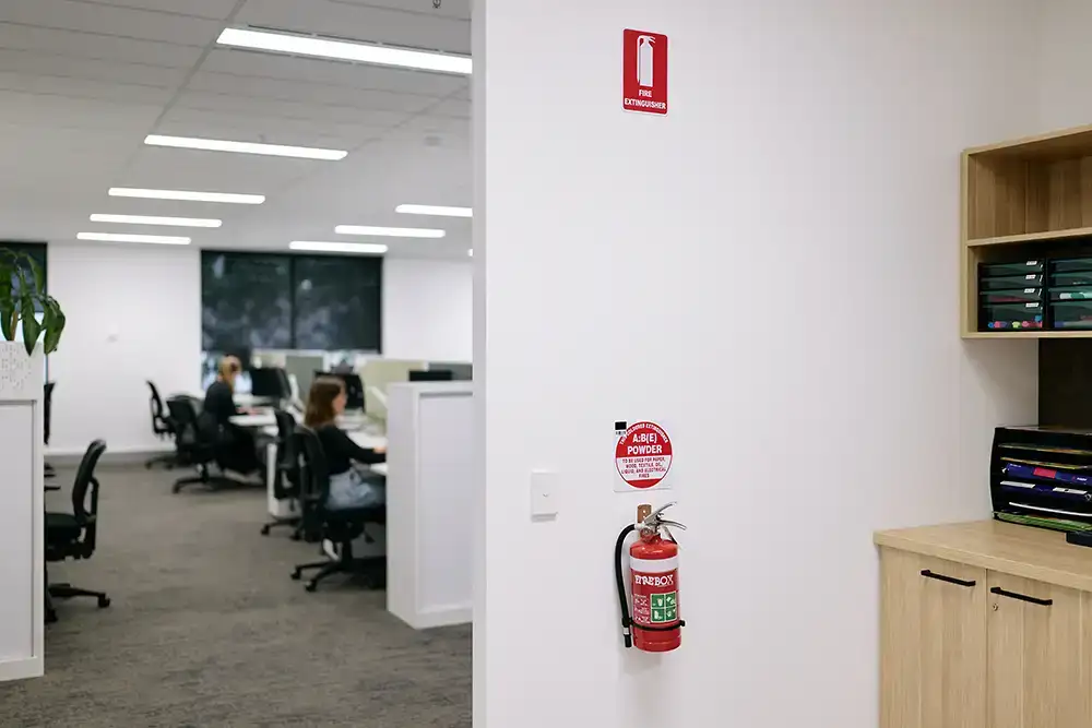 A dry chemical powder fire extinguisher mounted on a wall in an office utility room. Signs and labels indicating that there is an extinguisher here are clearly visible. Out of focus, in the background, people are working at desks in the office.