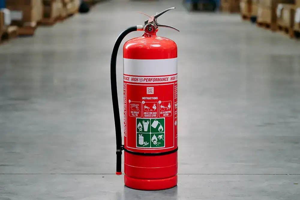 A red fire extinguisher with a white band around it sitting on a concrete floor in a warehouse. Instructions for use are clearly visible on the front of the extinguisher.