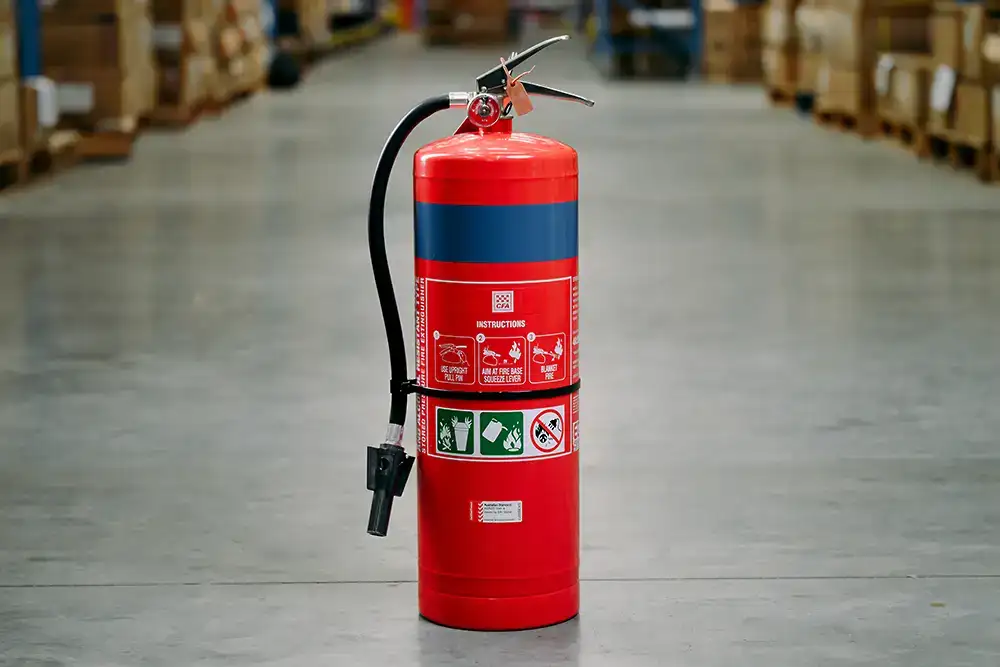 A red fire extinguisher with a blue band around it sitting on a concrete floor in a warehouse. Instructions for use are clearly visible on the front of the extinguisher.