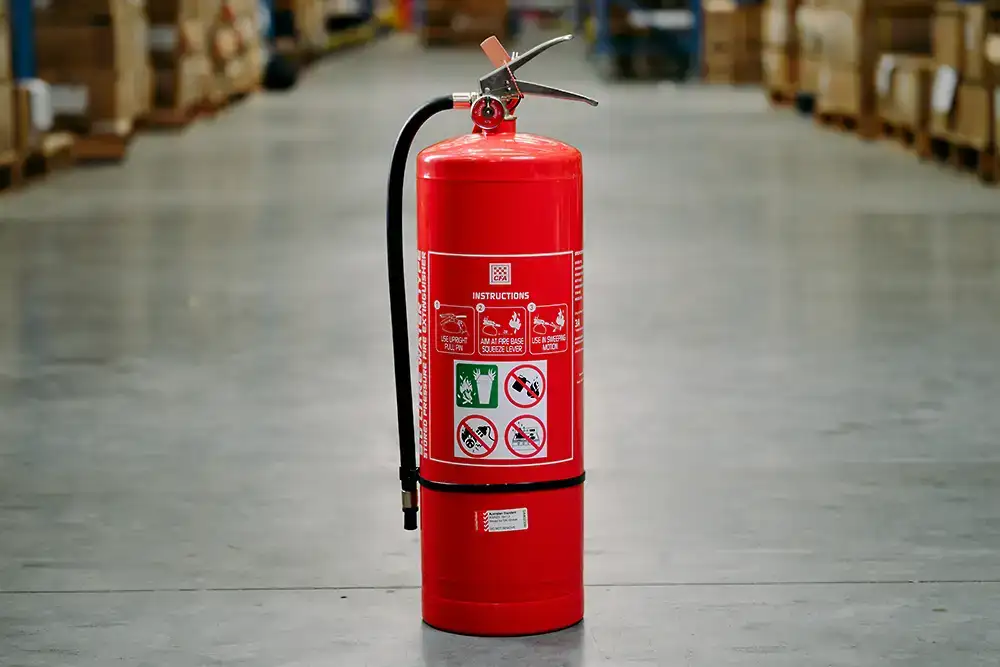A fire extinguisher which is red all over, sitting on a concrete floor in a warehouse. Instructions for use are clearly visible on the front of the extinguisher.