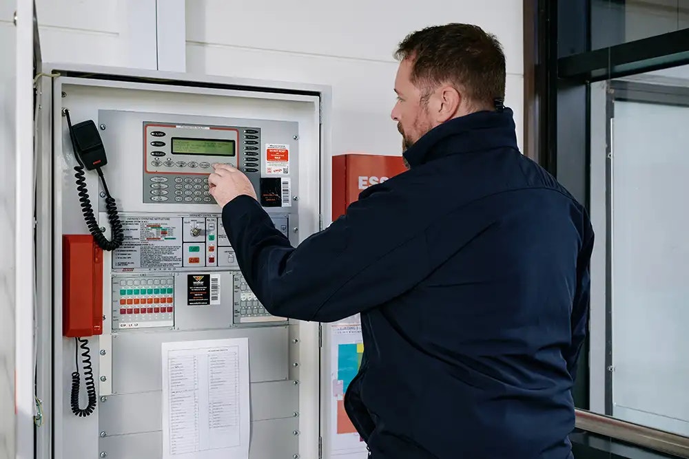 A person in navy work clothes standing in front of a fire alarm system control panel pressing a button.