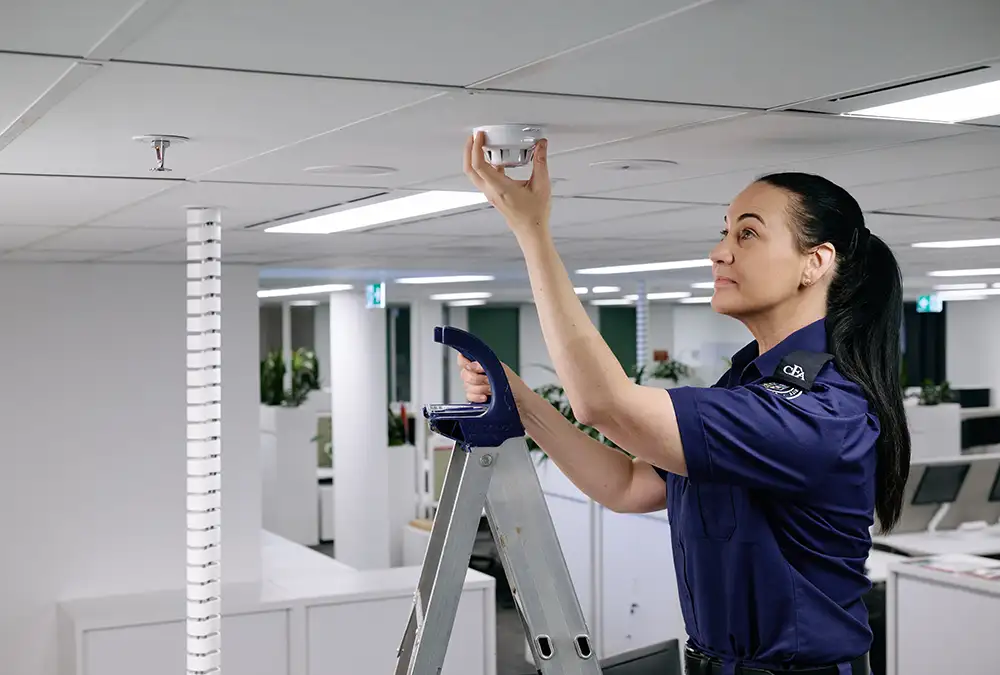 A fire equipment officer on a ladder checking a smoke alarm on a ceiling in an open plan office.