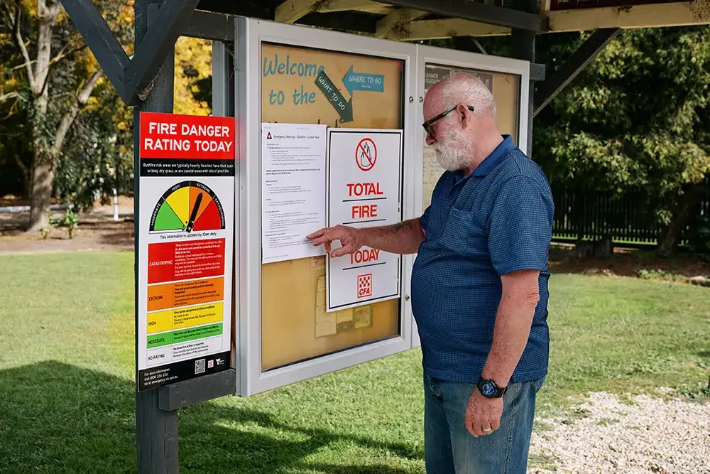 A man in a blue shirt checks a notice board in a caravan park. A Fire Danger Rating Today sign is set at extreme and a Total Fire Ban sign is displayed.