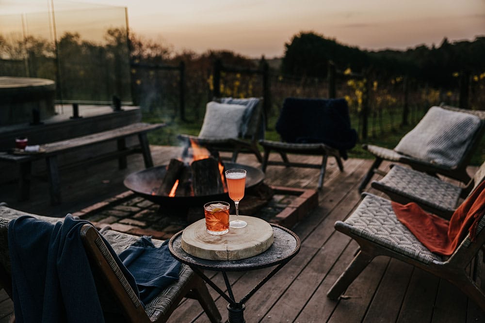 A fire pit on a raised brick platform on a wooden deck at sunset, surrounded by chairs and some drinks.