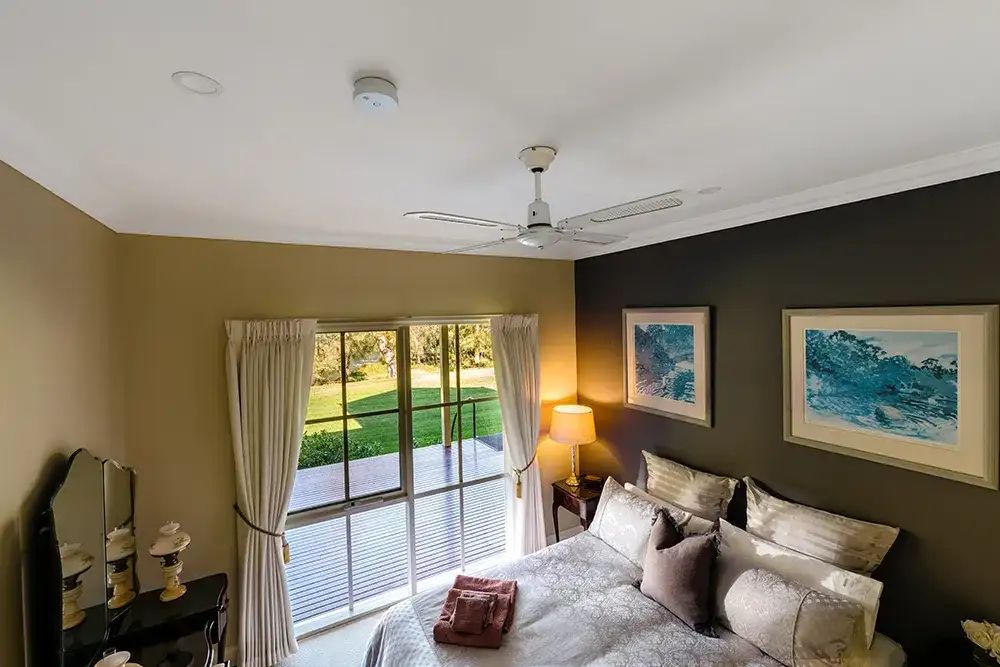 Modern heritage-style bedroom with a large window overlooking a garden. The room features a bed with plush pillows, warm lighting, a ceiling fan and a smoke alarm installed on the ceiling away from the fan.