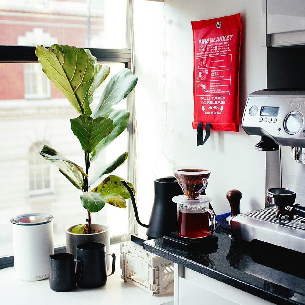 A fire blanket hanging on a wall in an apartment near a coffee machine, kettle, and a plant.