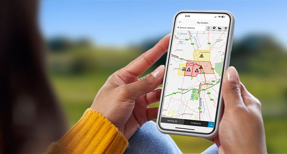 A person sitting outdoors in a beautiful country scene holding a phone which has a map and emergency icons on it.
