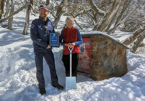 A person in a blue uniform holding a certificate and another person in civilian clothes holding a shovel next to a stone shed in the snow with snow gums behind.