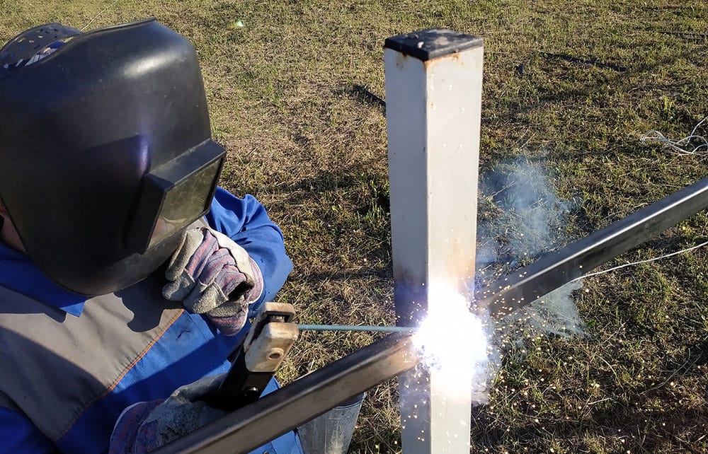 A person welding a metal fence outside. There are sparks coming from the activity falling towards the ground.