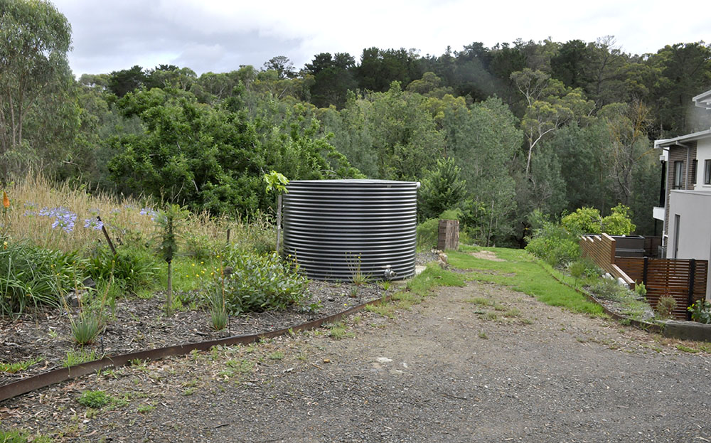 An above ground metal water tank with the compliant outlet located behind a house surrounded by bushland. The tank is located next to a gravel driveway with space for a fire truck to drive up next to the tank.
