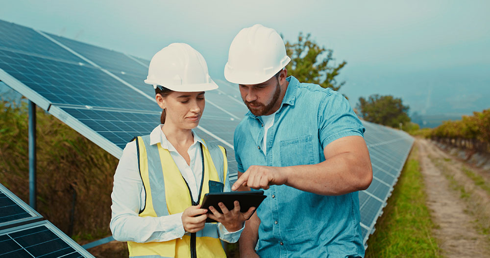 A consultant wearing a hard hat and a hi vis vest is discussing something on an iPad with a man in a hard hat. They are standing in front of solar panels in a field.
