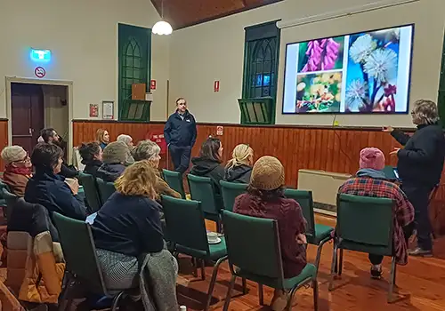 A group of people sitting in an old community hall listening intently to a presentation about ecology. Photos of plants and flowers are being displayed on a screen on the wall.