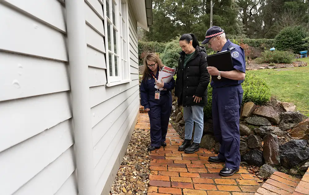 A male CFA member and female CFA member are standing with a female resident looking at a parthway made of rocks and bricks next to a house.