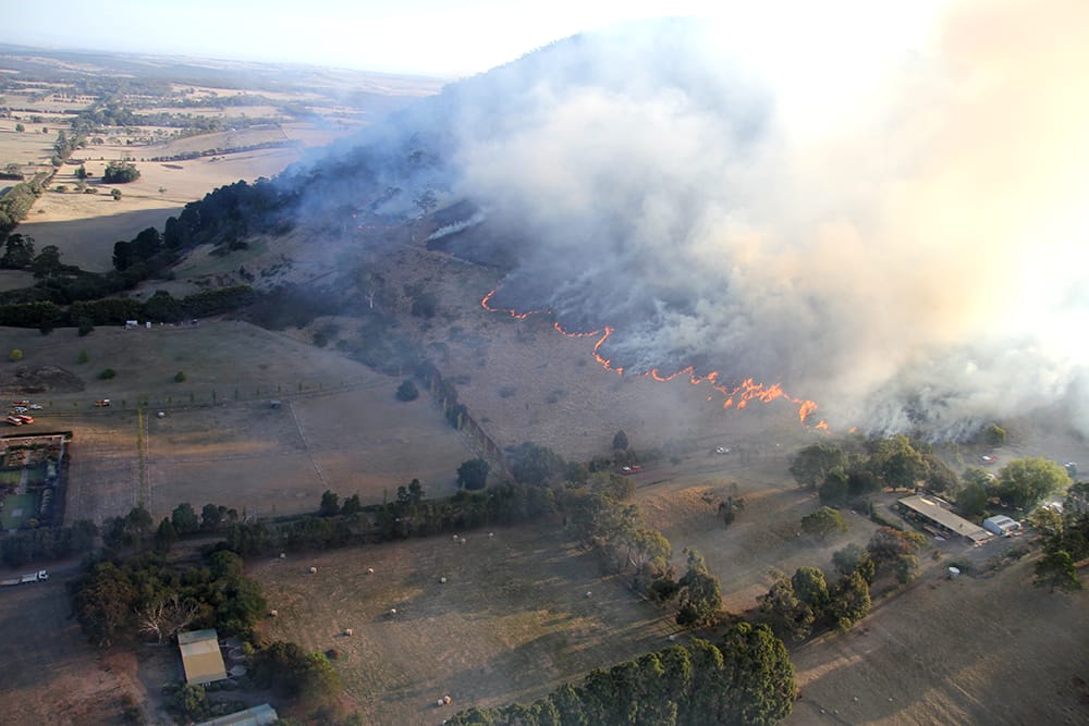 Aerial view of a grassfire spreading across a landscape and down a hillside toward property and homes, with smoke rising and flames. Some properties have hay bails.