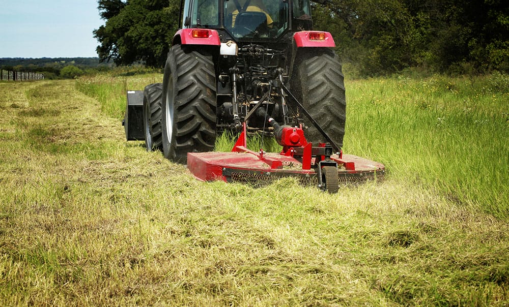 A red tractor slashing a grassy field with a rotary cutter attachment, leaving a path of cut grass behind it.