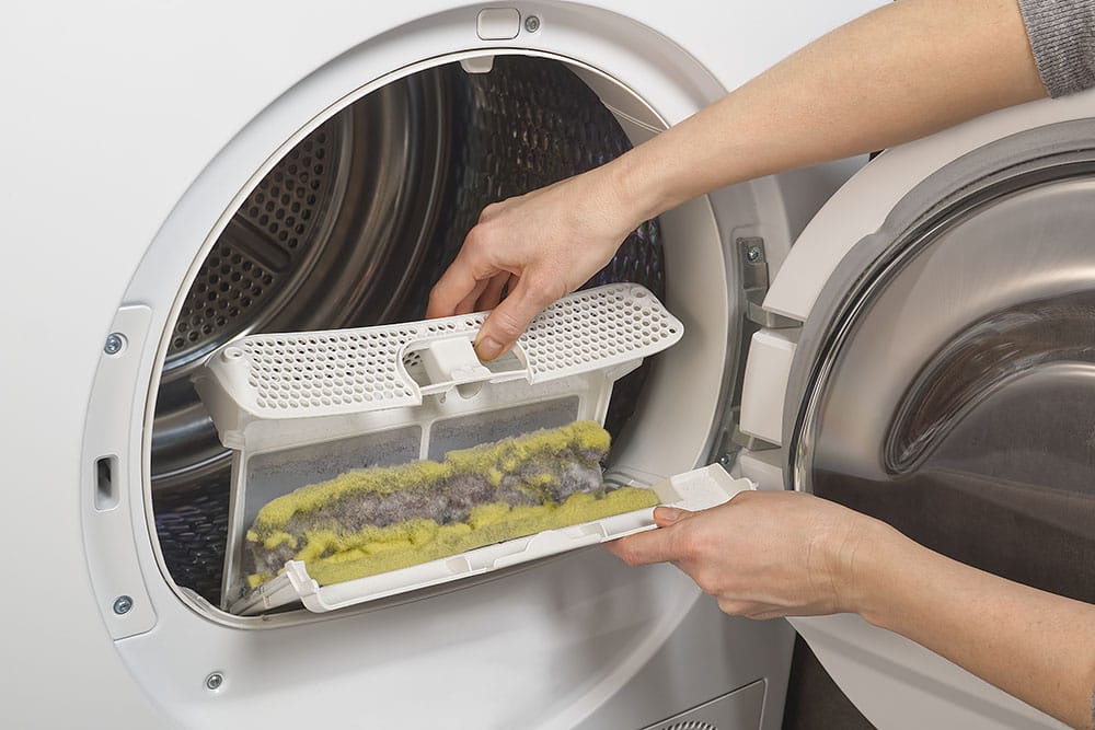 A person removing a lint filter from a dryer. The filter is filled with yellow and grey lint.