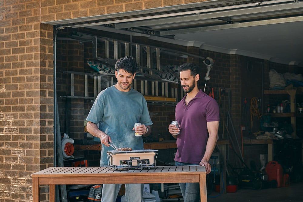 Two young men cooking on a hibachi coal BBQ outside the open door to a garage.