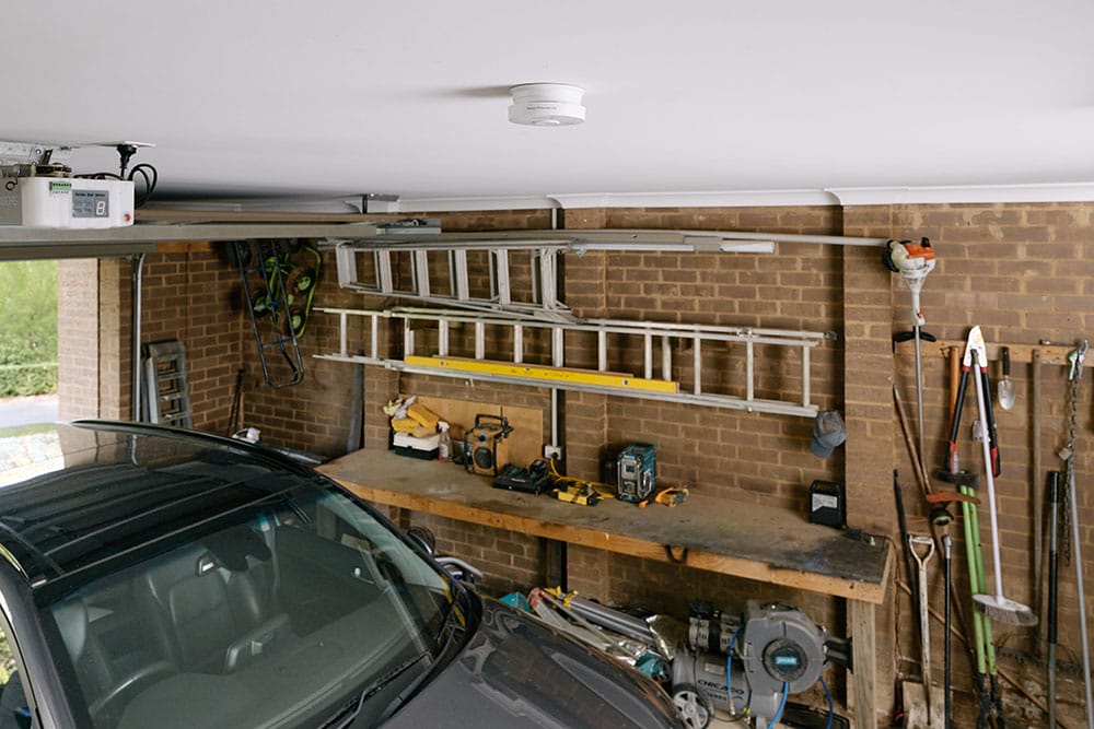 A smoke alarm on the ceiling in a garage with a car, work benches and items stored on walls.