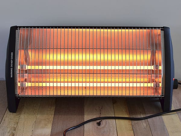 New electric heater turned on with glowing orange-red heating elements, placed on a wooden floor against a light-colored wall.