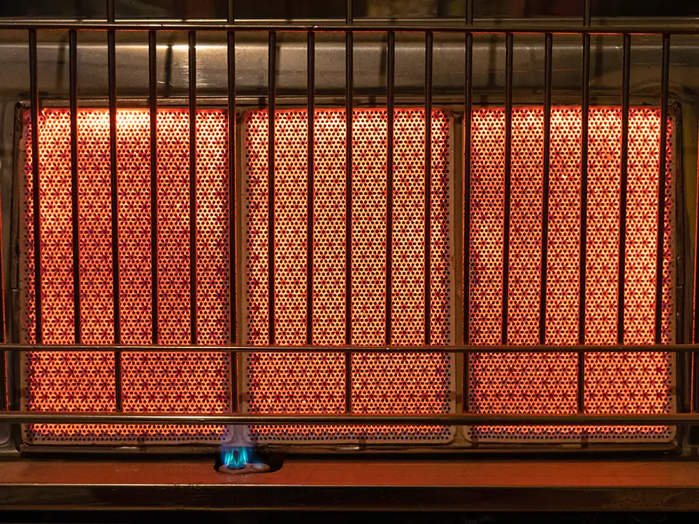 Three glowing red ceramic burner panels of a gas heater behind a protective metal grill, with a small blue pilot flame below