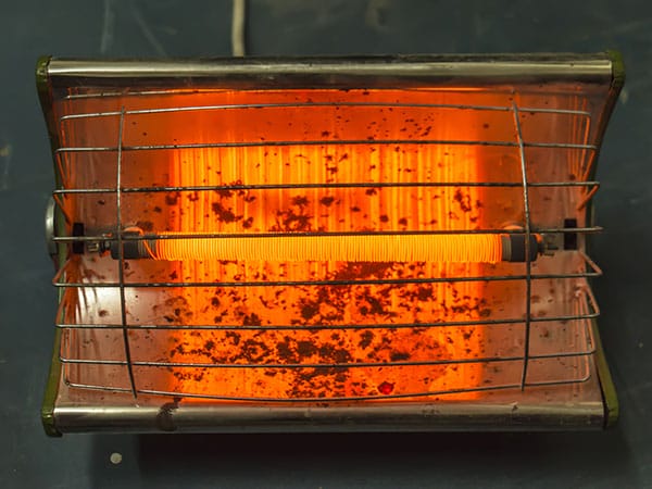 Old electric heater turned on with a glowing orange-red heating element, framed by a rusty reflector plate and a metal safety guard.