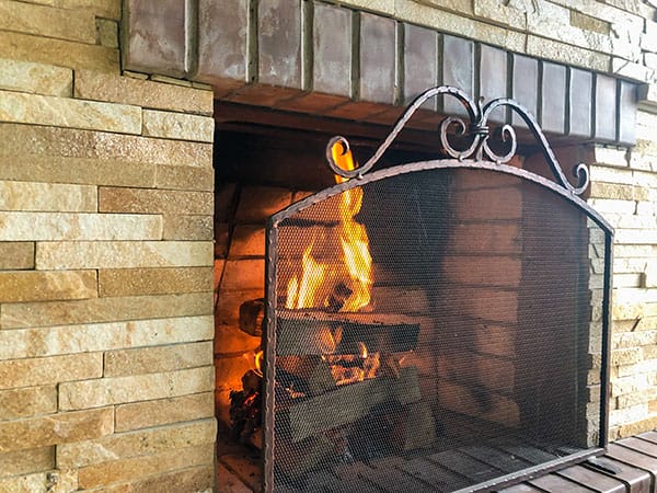 Beige brick fireplace with a roaring fire behind a decorative metal fire protective screen.
