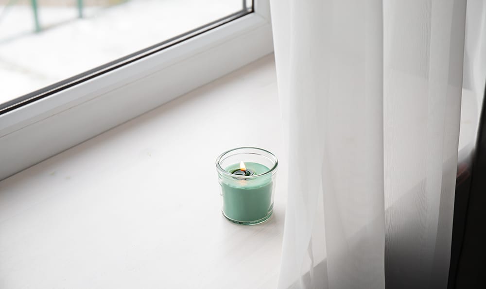A small green candle in a glass jar is lit on a windowsill next to sheer white curtains